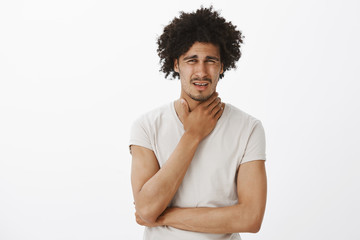 Guy feeling pain in throat, catching cold, frowning and grimacing from painful feeling, rubbing neck, standing intense over gray background, asking wife to make tea to end suffering