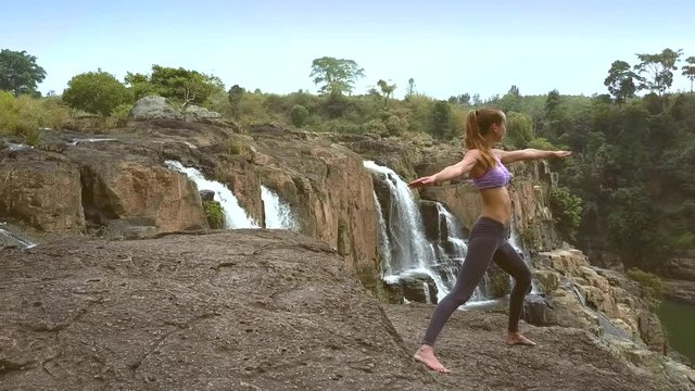 blond girl stands in yoga pose on rock near waterfall