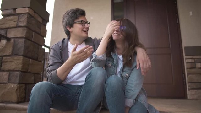 Smiling young guy in glasses is hugging his happy young wife sitting on their house porch. Family values concept. Slider slow motion medium shot