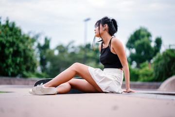 A very young and sophisticated Chinese Asian skater girl sitting and chilling on the ground while striking an alluring pose at a skate park.