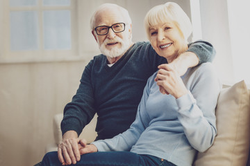 Happy marriage. Pleasant senior man hugging his wife while looking at you