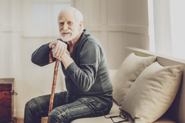 Pleasurable relaxation. Positive aged man holding his walking stick while resting at home