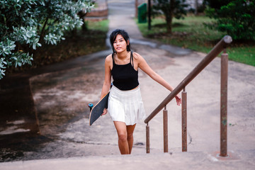 An attractive and cute Chinese Asian teenager walking at the park. She is carrying her skateboard and she is wearing a black tank top and white skirt.