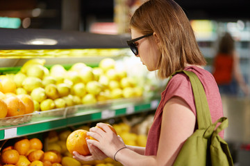 Obraz premium Photo of female teenager stands back, carries rucksack on back, chooses oranges for making fruit salad, wears shades, keeps to healthy diet, poses in supermerket. People and purchasing concept