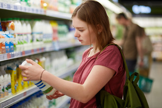 Photo of attractive young female consumer model with bobbed hairstyle, dressed in casual t shirt, stands in big store, holds bottle of milk, focused on label, wants to buy some dairy products