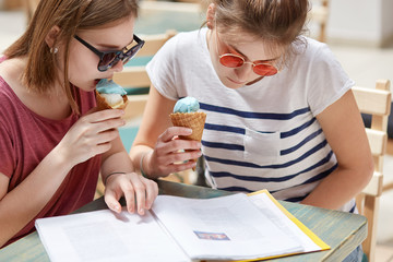 Cropped shot of serious focused young females look attentively into menu of cafeteria, eat cold fruit ice cream, recreat during summer time together, wear sunglasses. What can we order there?