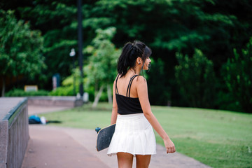 A portrait of a young, attractive, and too-cool-for-school Chinese Asian skater girl strolling in the park with her skateboard.