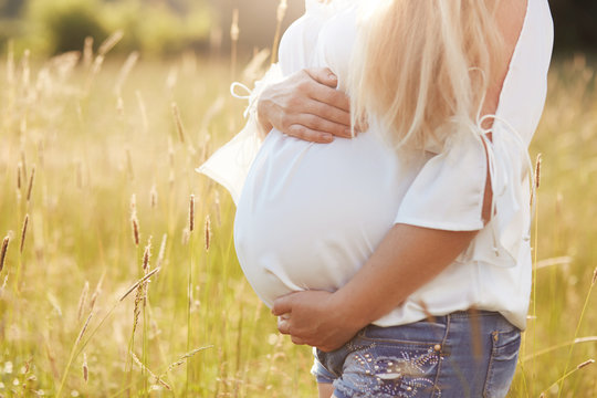 Cropped shot of unrecognizable young future mother being pregnant, keeps hands on tummy, stands on green field, wears white blouse enjoys sunshine and summer time. Motherhood and parenthood concept