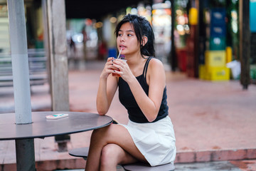 An attractive young Chinese Asian girl sitting at a hawker center while enjoying her cold beverage.