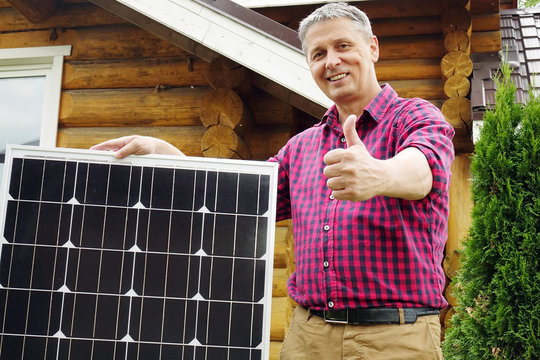 Solar panels in the hands of men . Energy production technologies. Wooden house background. The thumb of a person's hand upwards .