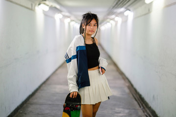 A studio portrait of a young, cute, cool and attractive Chinese Asian millennial girl holding her skateboard while posing glamorously. She is wearing trendy sports wear.