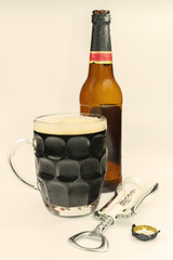 a glass mug of dark beer with a frothy cap, a beer bottle next to it, a metal lid and an opener