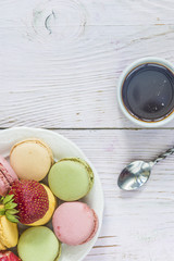 Coffee break with french macaroons/One strawberry on pastel colored french macaroons on a white plate and cup of black coffee on white paint wooden background 