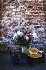 Travel inspiration - bunch of peony flowers, vintage suitcase and straw hat in loft style interior