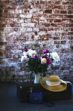 Travel inspiration - bunch of peony flowers, vintage suitcase and straw hat in loft style interior
