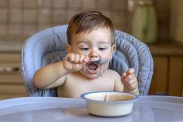 Funny little baby boy holding spoon and eating porridge trying to catch falling food with open mouth, learning to eat by itself