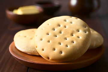 Traditional Chilean Hallulla bread rolls on wooden plate, butter, jam and cup in the back, photographed with natural light (Selective Focus, Focus through the middle of the roll)