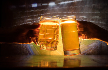 Two friends toasting (clinking) with glasses of light beer at the pub. Beautiful background with blurred view of playing game at the stadium.