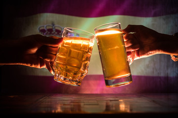 Two friends toasting (clinking) with glasses of light beer at the pub. Beautiful background with blurred view of flag of Spain. Support your country with beer concept.