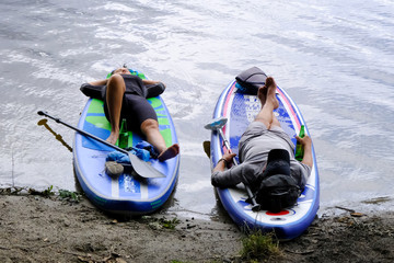 Fototapeta Letni wypoczynek nad wodą - kobieta i mężczyzna odpoczywający na brzegu jeziora na deskach z wiosłem, SUP, Stand Up Paddle obraz