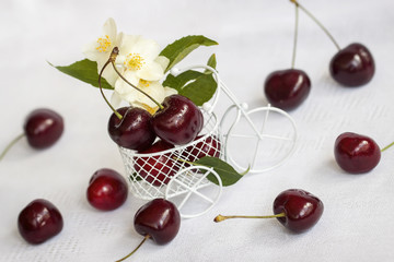 Small toy shopping cart, trolley closeup with ripe cherry, jasmine flowers on light background. Vegetarian concept, diet, detox, organic vitamins