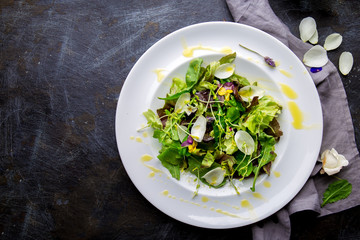 Healthy lettuce arugula salad with edible flowers and microgreens on white plate, dark background. Top view