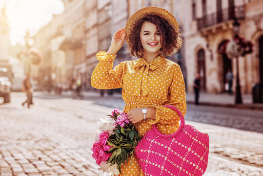 Outdoor portrait of young beautiful happy smiling girl wearing yellow polka dot dress, hat, holding straw bag with peonies, posing in street of european city. Spring, summer fashion. Copy space 
