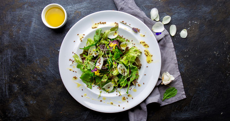 Healthy lettuce arugula salad with edible flowers, microgreens and linseeds on white plate, dark background. Top view