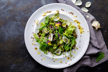 Healthy lettuce arugula salad with edible flowers, microgreens and linseeds on white plate, dark background. Top view