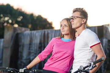 Fototapeta na wymiar Sport Concepts. Young Caucasian Couple with Mountain Bikes Posing Together Outdoors In Front of Waterfall on The Background.