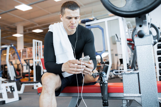 Portrait of handsome muscular man with prosthetic leg sitting on bench and relaxing listening to music from smartphone after working out in modern gym, copy space - Powered by Adobe