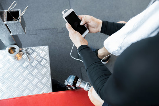 High angle portrait of unrecognizable muscular man with prosthetic leg holding smartphone sitting on bench and listening to music after working out in gym, copy space