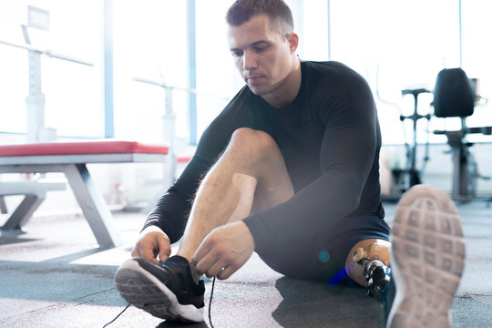Full length portrait of muscular sportsman with prosthetic leg tying shoe sitting on floor in modern gym, copy space - Powered by Adobe