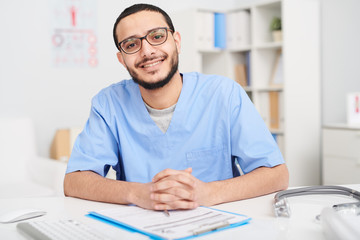 Obraz na płótnie Canvas Portrait of smiling young Middle-Eastern doctor wearing glasses sitting at desk in office and looking at camera