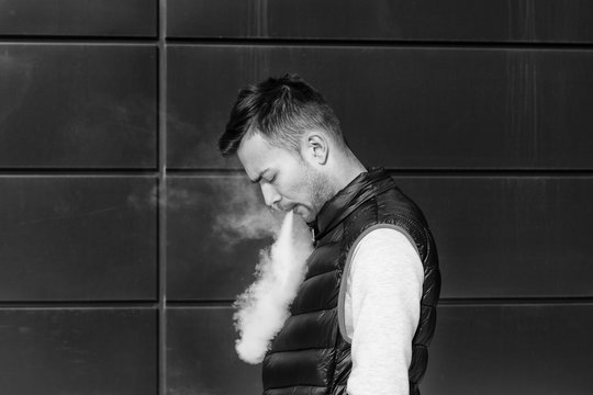 Vape man. Portrait of a handsome young white guy in casual clothes vaping an electronic cigarette opposite the modern background.