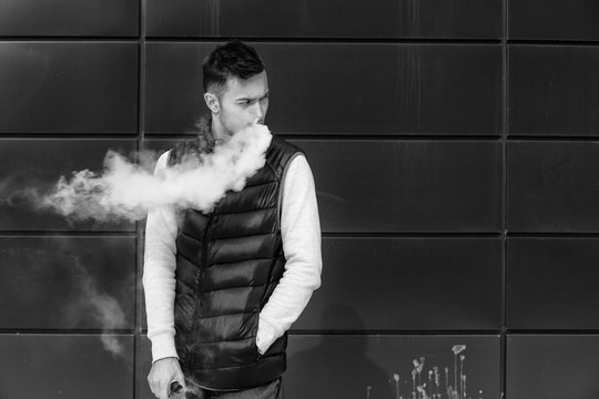 Vape man. Portrait of a handsome young white guy in casual clothes vaping an electronic cigarette opposite the modern background.