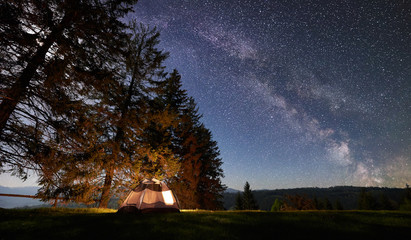 Night camping in mountains. Tourist tent by brightly burning campfire under beautiful dark blue starry sky, Milky way. High pine trees on background. Beauty of nature, travelling and tourism concept.