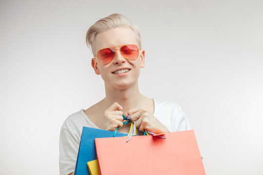 Male fashion, shopping concept. Portrait of blonde young man with pink sun glasses, posing with coloured shopping bags over studio white background. Shopping Moll, Customers, Trade