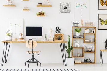 Real photo of a simple home office interior with a desk, computer, chair, cupboard, cactus, posters with dragonflies and butterflies on the wall. Place your product on the empty screen