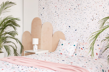 Pink blanket on patterned bed in bright bedroom interior with plant and wallpaper. Real photo