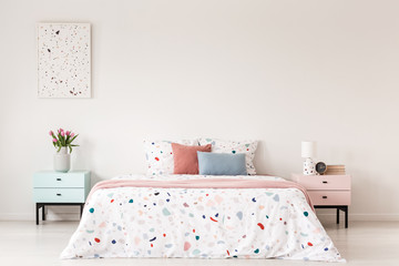 Poster above cabinet with flowers next to bed with colorful pillows in white bedroom interior. Real photo - obrazy, fototapety, plakaty