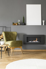 Mockup of white empty poster above fireplace in grey flat interior with green armchair. Real photo