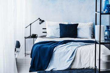 Navy blue blanket on bed in pastel bedroom interior with lamp on table - obrazy, fototapety, plakaty