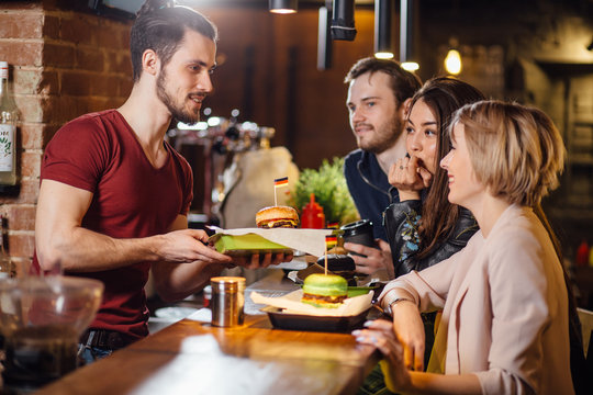 Group of happy smiling friends ordering food through barkeeper at the counter in modern restaurant with loft style, bricks and pipes, interior,