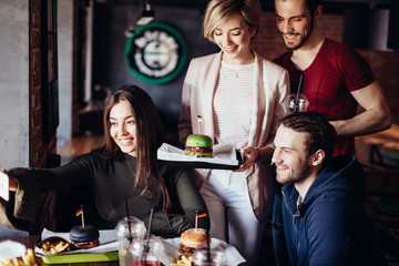 Group of positive cheerful friends having fun, taking selfie with smartphone in burger bar. Young blonde woman holding tray with ready green burger