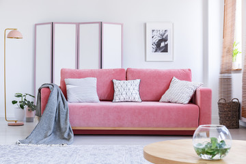 Real photo of a pink couch with white pillows and a blanket standing in a cozy living room interior with a table with a vase, a lamp and a screen