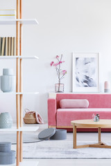 Real photo of a pink sofa with a cushion standing behind a wooden table and in front of a shelf with a poster and flowers in living room interior with a shelf with ornaments in the front