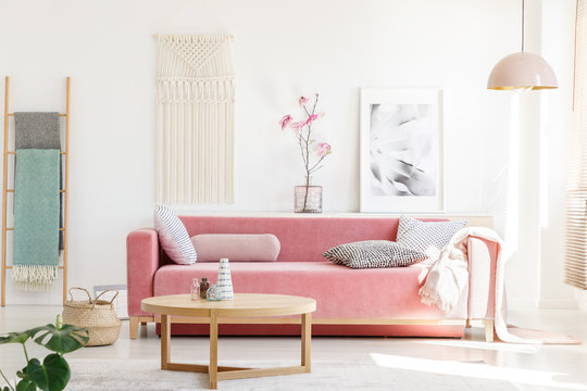 Real photo of a pink sofa with cushions and blanket standing behind a wooden table in bright living room interior with a hanging lamp, ladder, poster and flowers
