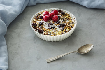 Muesli, raspberries and raisins  in a bowl next to a spoon on a grey table