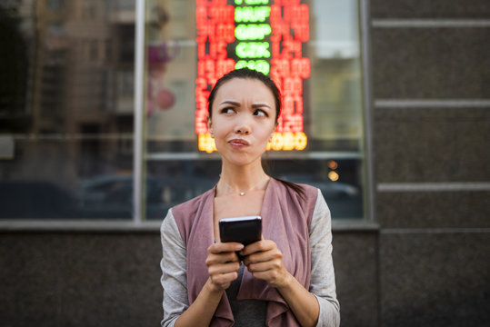 A young beautiful asian woman using an application in her smart phone to check currency exchange rates in front of an illuminated information board. Communicate about money value, international